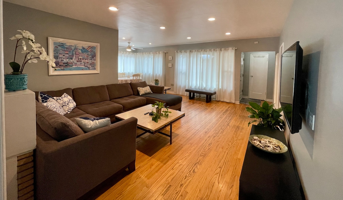 A spacious living area is furnished with a large sectional sofa and a coffee table, featuring greenery and decorative items. Natural light fills the room through sheer curtains, creating a bright atmosphere. A dining area with a table is visible in the background, enhancing the homey feel.