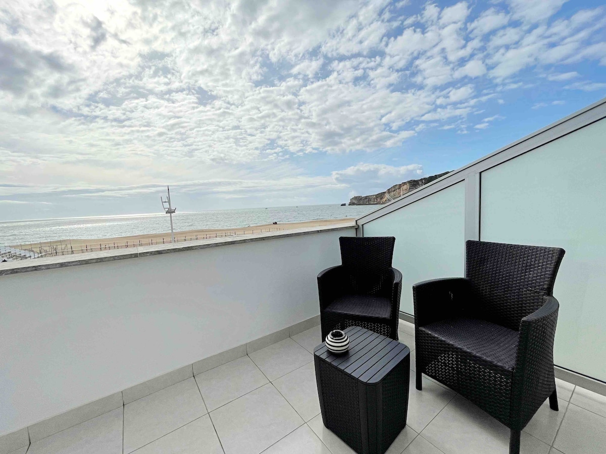 A private balcony features two black wicker chairs and a small table, with a view overlooking the beach and the Atlantic Ocean. The sky is partly cloudy, creating a serene atmosphere. The balcony surfaces are tiled, enhancing the modern design of the apartment.