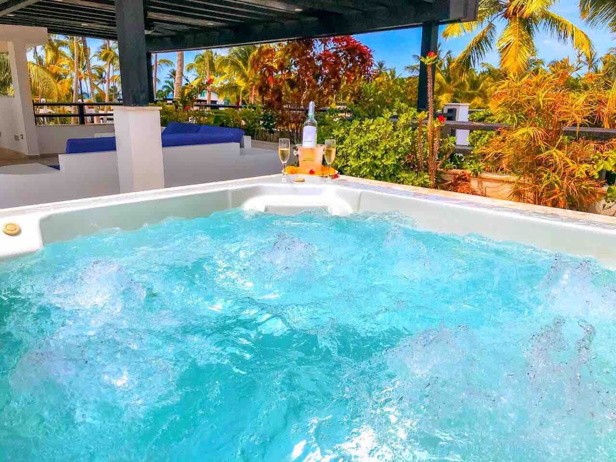 A private hot tub is visible, filled with clear bubbling water. Refreshing drinks and glasses are placed on a small tray. Lush greenery and palm trees surround the area, creating a tropical atmosphere. The blue sky can be seen above, enhancing the serene environment.