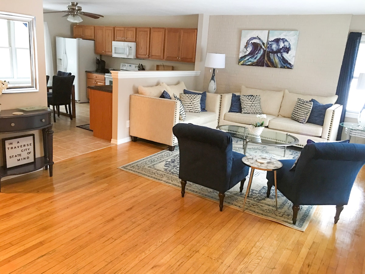 An open living area features comfortable seating arrangements, including two blue chairs and a beige sofa adorned with textured pillows. A coffee table is centered on a patterned area rug over hardwood floors. The kitchen can be seen in the background, equipped with white appliances and wooden cabinetry.