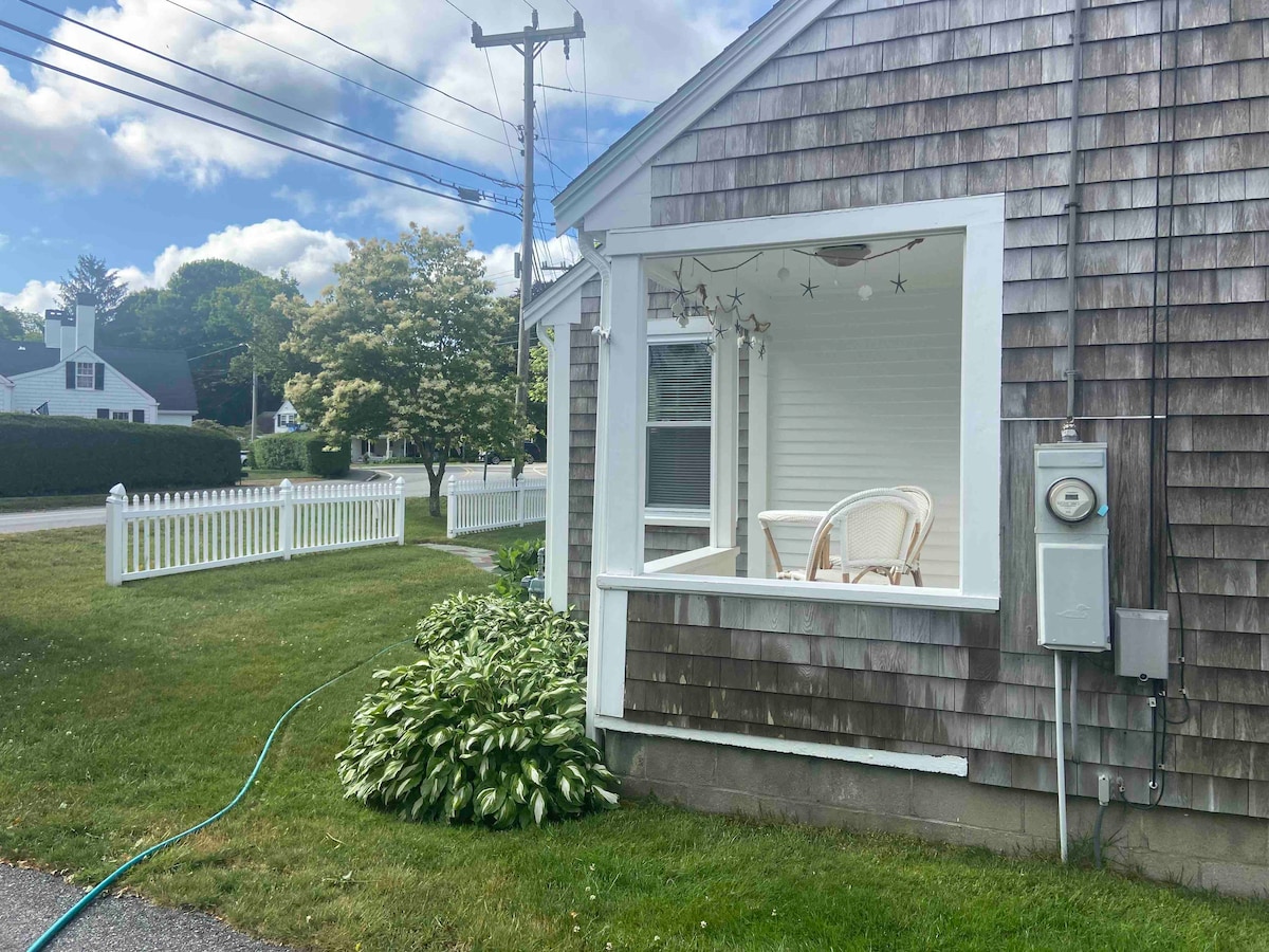 The exterior view showcases a cottage with a window framed by white trim, revealing a small seating area inside. Lush greenery and a white picket fence outline the yard, while power lines run above the scene. The sky is partly cloudy.