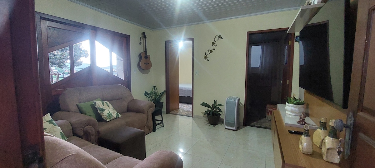 The living area features comfortable seating on plush sofas, a decorative plant placed nearby, and a wall-mounted television. Natural light enters through large windows, and a guitar is hanging on the wall, adding a homey touch to the space.
