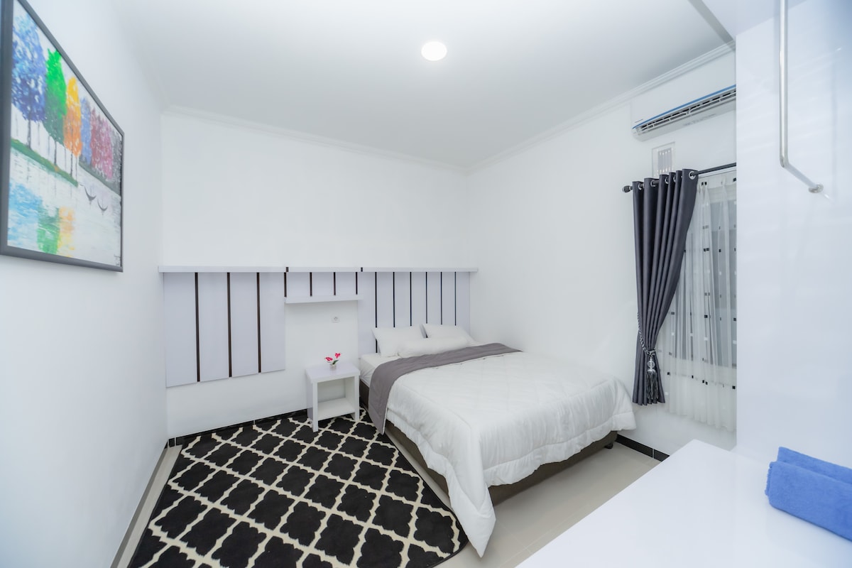 A bright sleeping area features a spacious bed with neatly arranged bedding. A decorative painting adorns the wall, while a small bedside table holds a flower vase. Curtains frame a window, and a patterned rug covers the floor, adding texture to the room.
