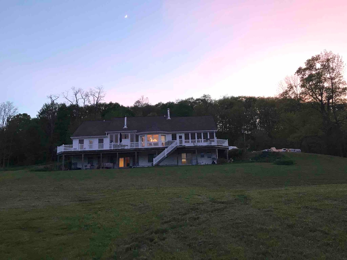 A spacious two-story home is set against a backdrop of a vibrant sunset. The structure features an expansive deck and numerous windows that reflect the warm evening light. Surrounding greenery enhances the tranquil setting, providing a serene atmosphere.