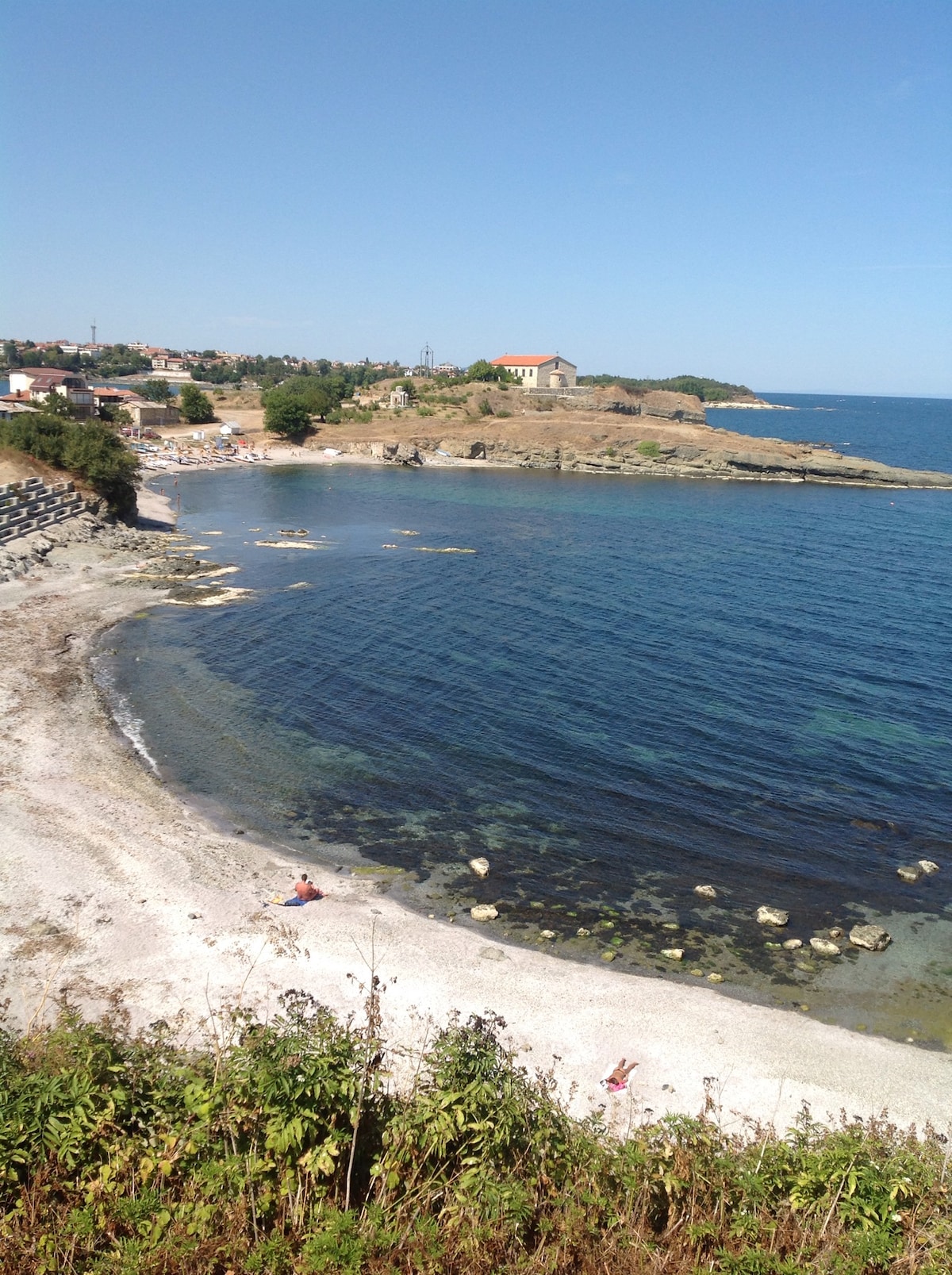 A tranquil coastline is visible, highlighted by clear blue water lapping against a pebbled shore. Soft white sand is interspersed with small rocks. In the background, a gentle rise features a few buildings, framed by greenery and distant hills under a clear sky.