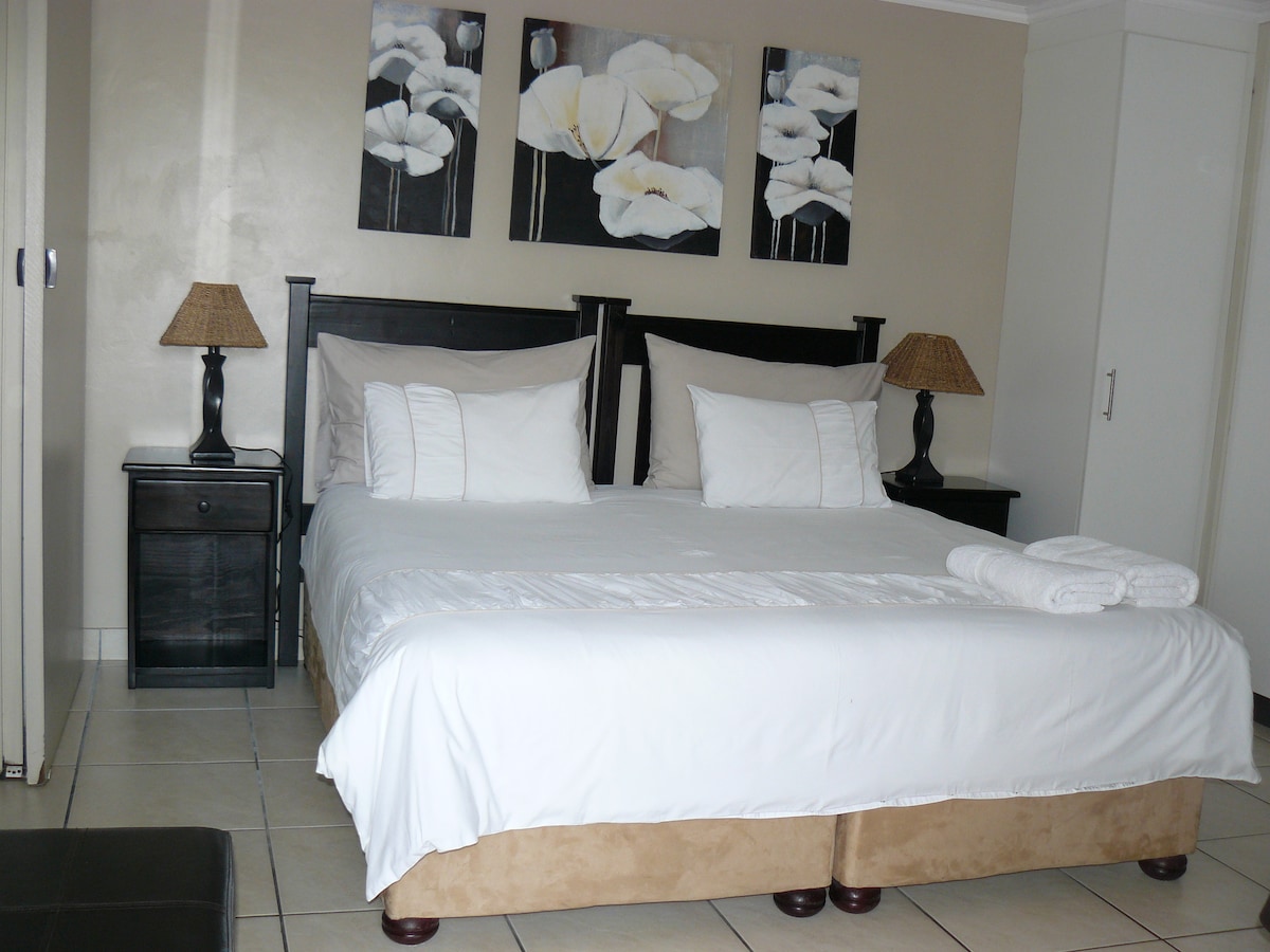A spacious room features a comfortable king-size or twin beds with soft linens. Two bedside tables, each with a lamp, flank the bed. Wall art displaying floral designs adds a decorative touch. A dresser is visible in the background against a light-toned wall.