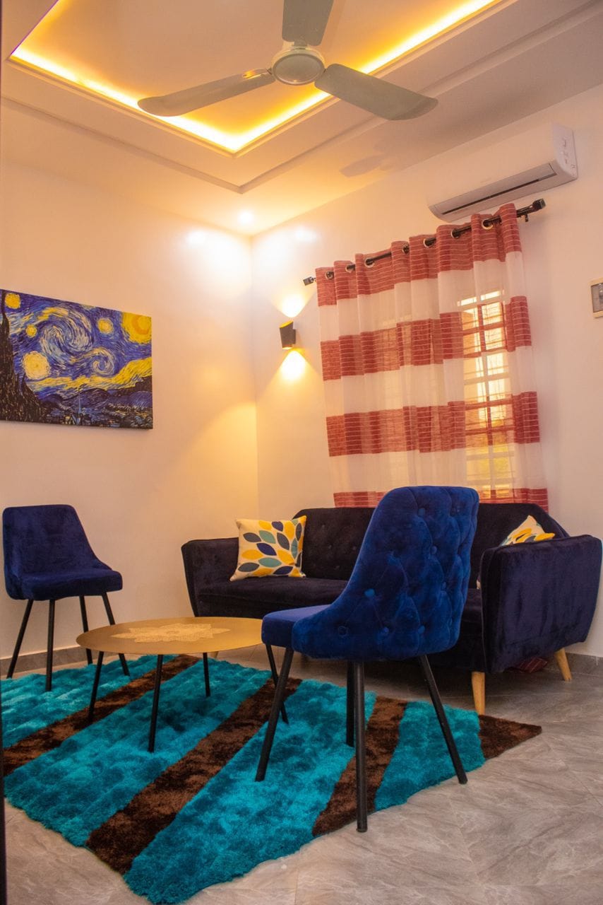 A cozy living area features a deep blue sofa paired with two matching chairs. A round coffee table is centered on a textured area rug in various shades of blue and brown. Artistic wall decor and elegant lighting add to the inviting ambiance.