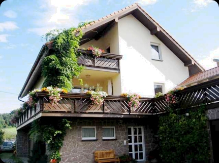 The exterior features a three-story building adorned with greenery and colorful flower boxes. A covered balcony extends from the upper level, offering a space for relaxation. A wooden bench is placed near the entrance, surrounded by well-maintained landscaping.