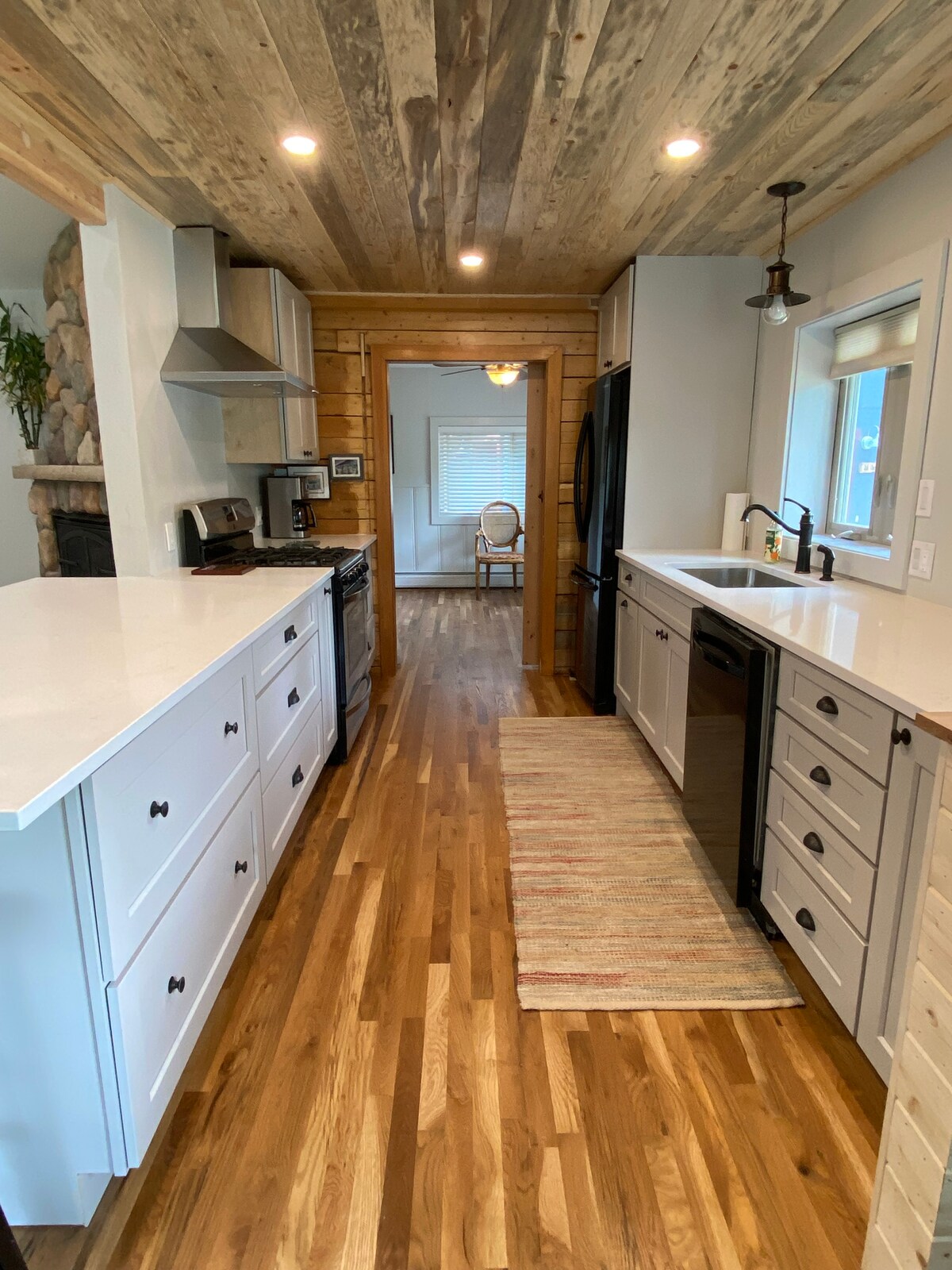 The kitchen features a combination of light and dark cabinetry with a central island, offering ample counter space. Wooden beams accentuate the ceiling, and a stylish rug runs the length of the space. Modern appliances are arranged alongside windows, providing natural light.