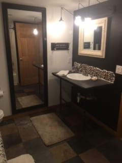 A bathroom space features a large mirror above a console sink, with decorative tiles outlining the backsplash. Soft lighting from overhead fixtures complements the dark accent wall. A comfortable rug lies beneath the sink area, and a wooden door adds warmth to the modern design.