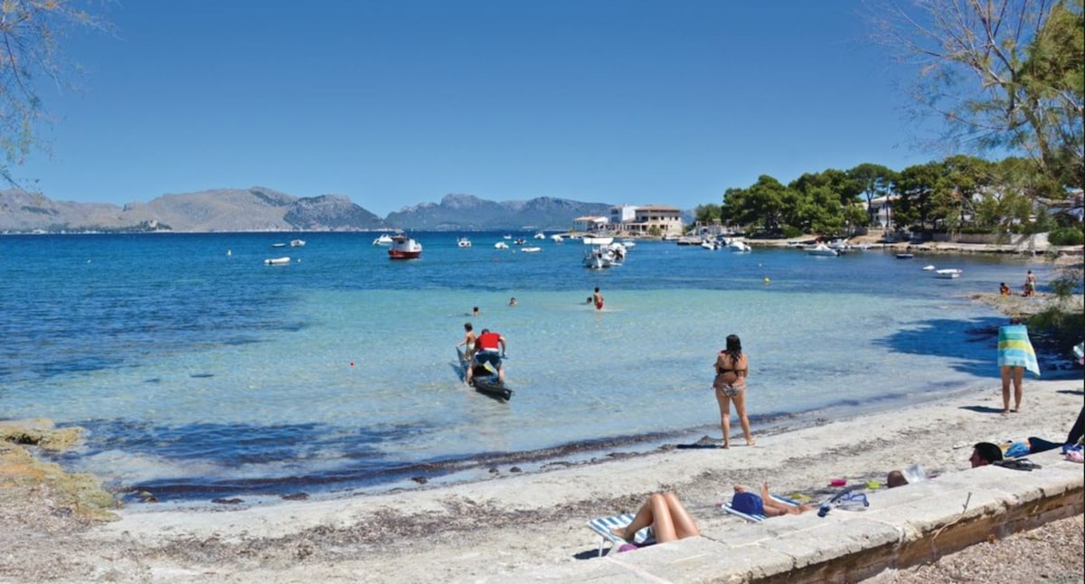 A sandy beach with clear water is shown, featuring people enjoying the sun and swimming. Small boats are anchored nearby, and the distant mountains are visible under a clear blue sky. Trees line the beach area, providing some shade.