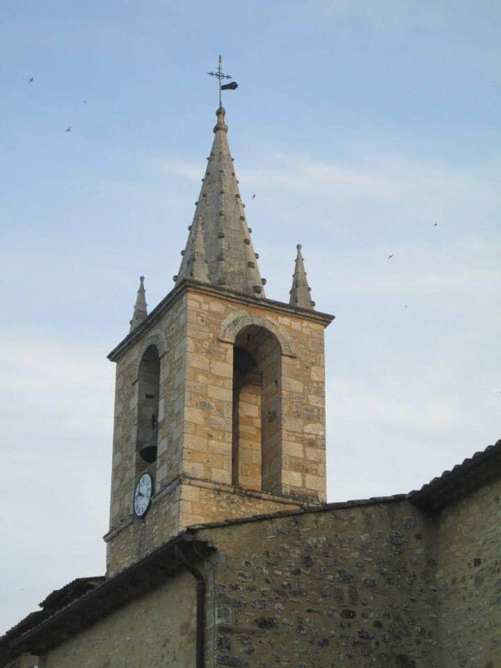 A stone church tower rises into the sky, featuring a tall spire topped with a weather vane. The clock face is visible on one side of the tower, while the textured stone walls reflect a historical architectural style. Subtle details of clouds are present in the background.