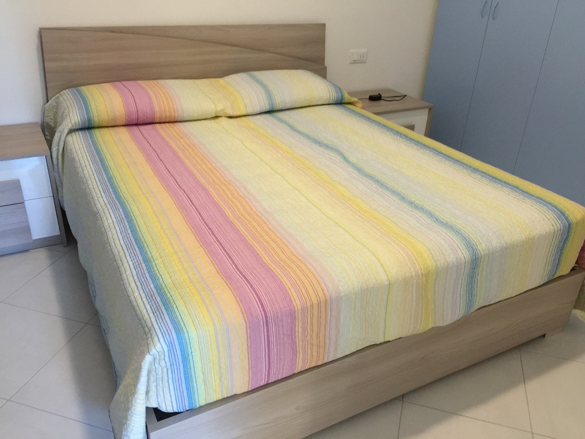 A spacious bed is centered in the room, featuring a multi-striped coverlet in pastel colors. Flanking the bed are two bedside tables, one with a small remote control, while a large wardrobe with sliding doors is visible in the background.