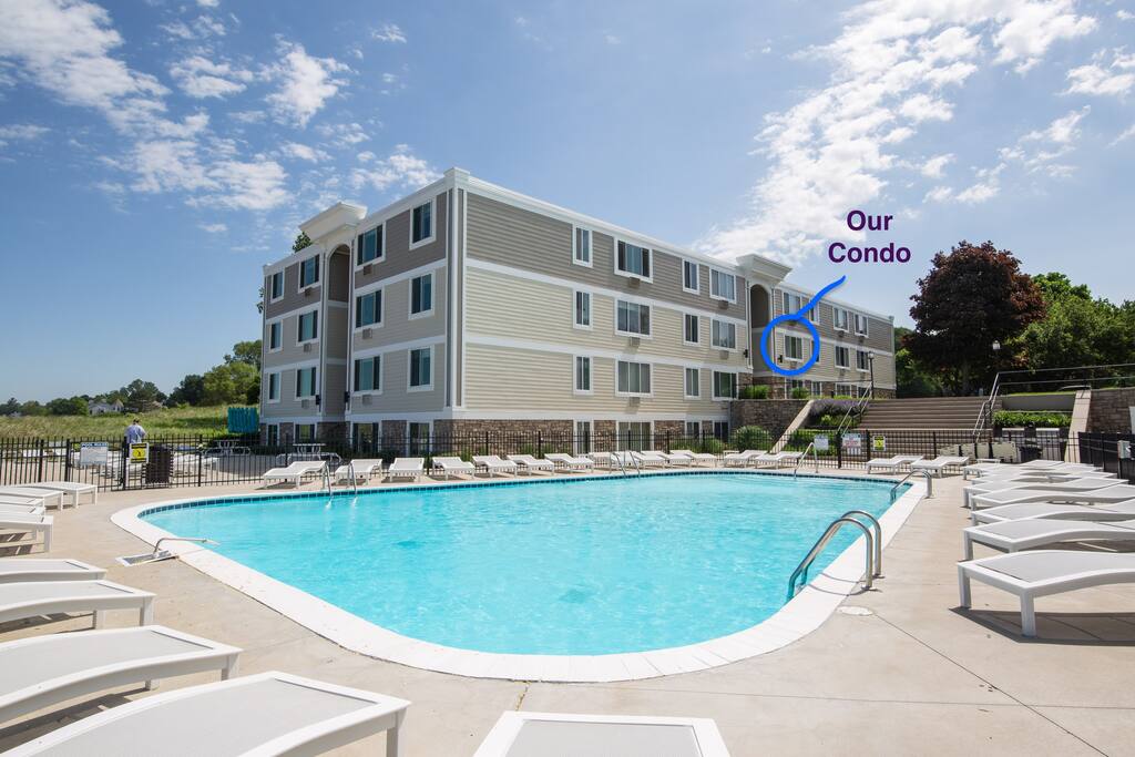 A spacious outdoor pool is surrounded by lounge chairs, inviting relaxation. The three-story condo building is visible in the background, marked for easy identification. Lush greenery and blue sky enhance the setting, creating a welcoming environment for guests.