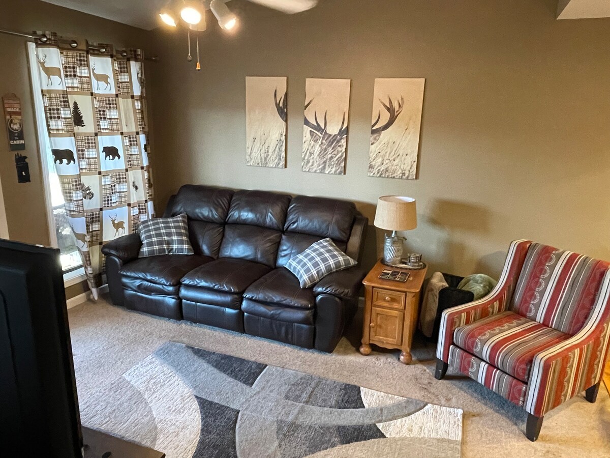 A comfortable living area features a dark leather couch and a striped armchair, with decorative throw pillows accenting the seating. Three nature-themed wall art pieces are displayed above the couch. Soft lighting and a small wooden side table enhance the cozy environment.