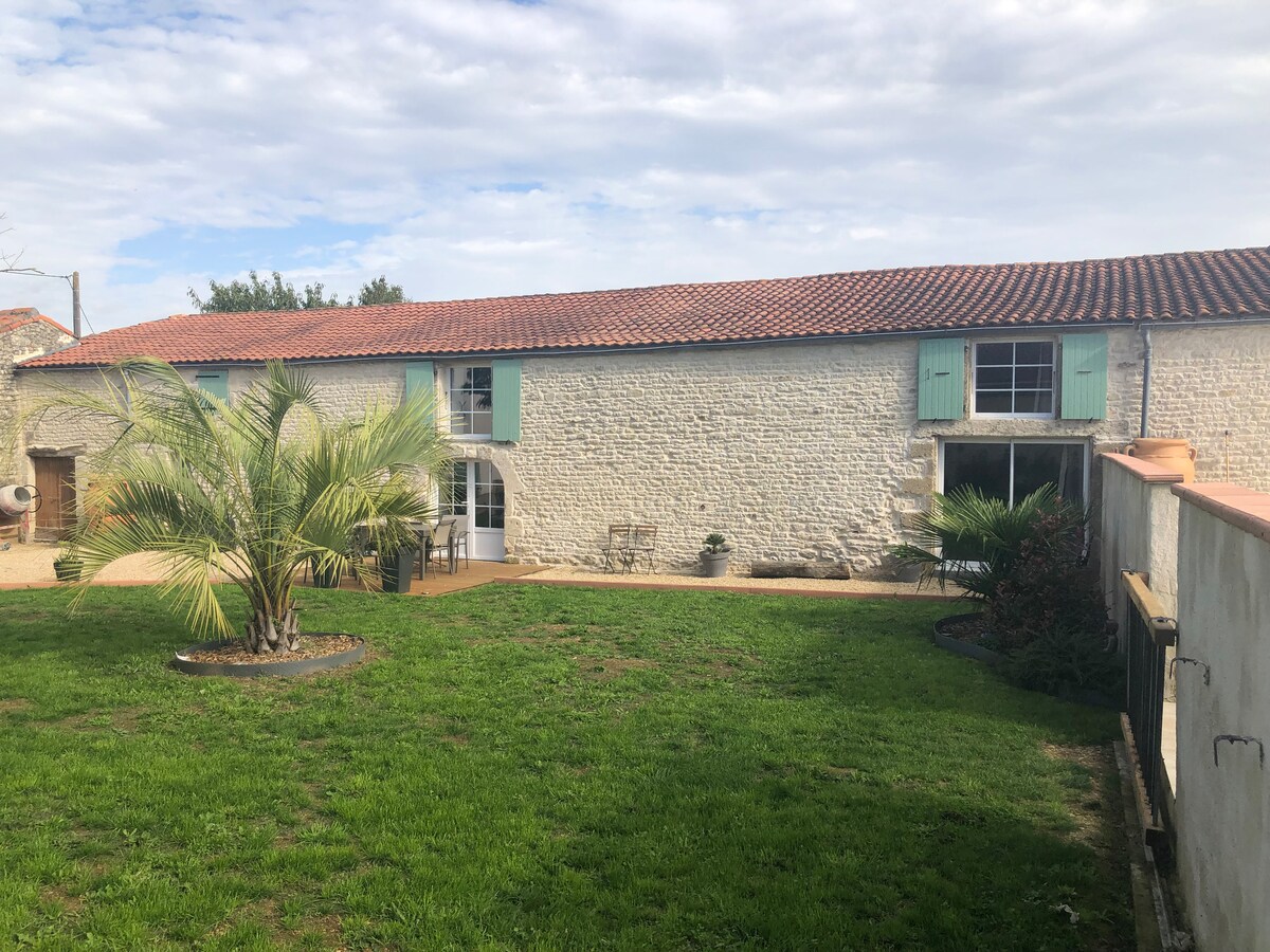 The exterior of the renovated barn features a grassy area with a central palm tree. Stone walls are complemented by green shutters and a red-tiled roof. A patio with chairs is visible, along with garden elements enhancing the tranquil setting.