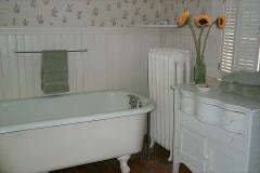 A spacious bathroom features an original claw foot tub, complemented by a vintage white radiator and a small cabinet. Soft green towels are neatly arranged, and a vase with sunflowers provides a subtle pop of color. Decorative wallpaper adds a charming touch.