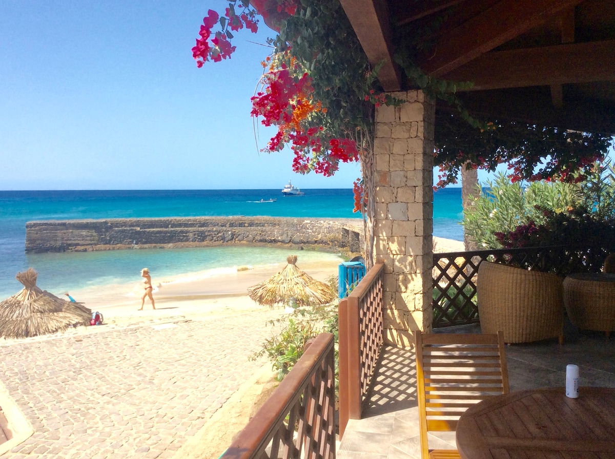 A view from the bungalow's terrace showcases a sandy beach leading to tranquil blue waters. Natural elements like wooden furniture and vibrant flowers provide a warm touch. Straw umbrellas offer shade, while a few guests can be seen enjoying the beach in the distance.