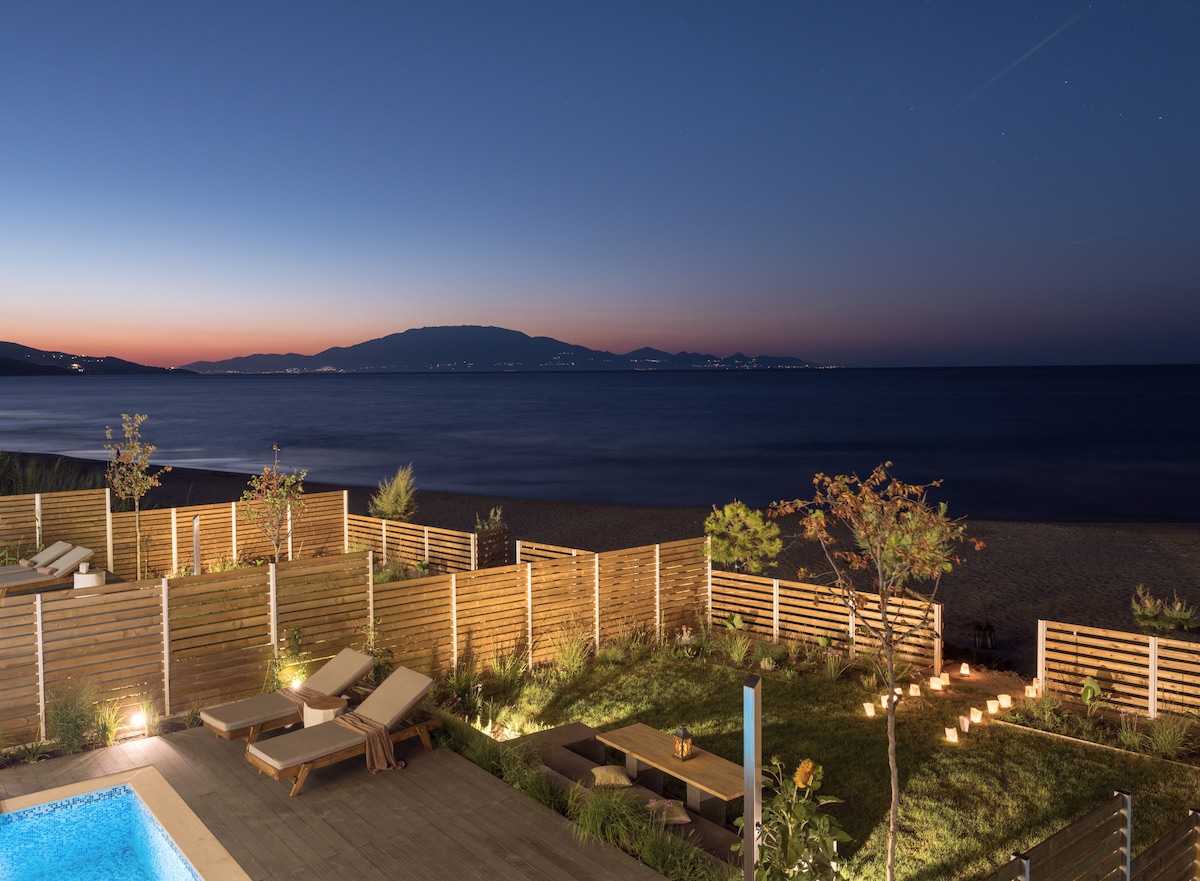 A serene view from the property overlooks the calm sea at twilight, with silhouettes of distant mountains visible. A swimming pool is in the foreground, surrounded by sun loungers and landscaped gardens that include low wooden fencing and subtle lighting.