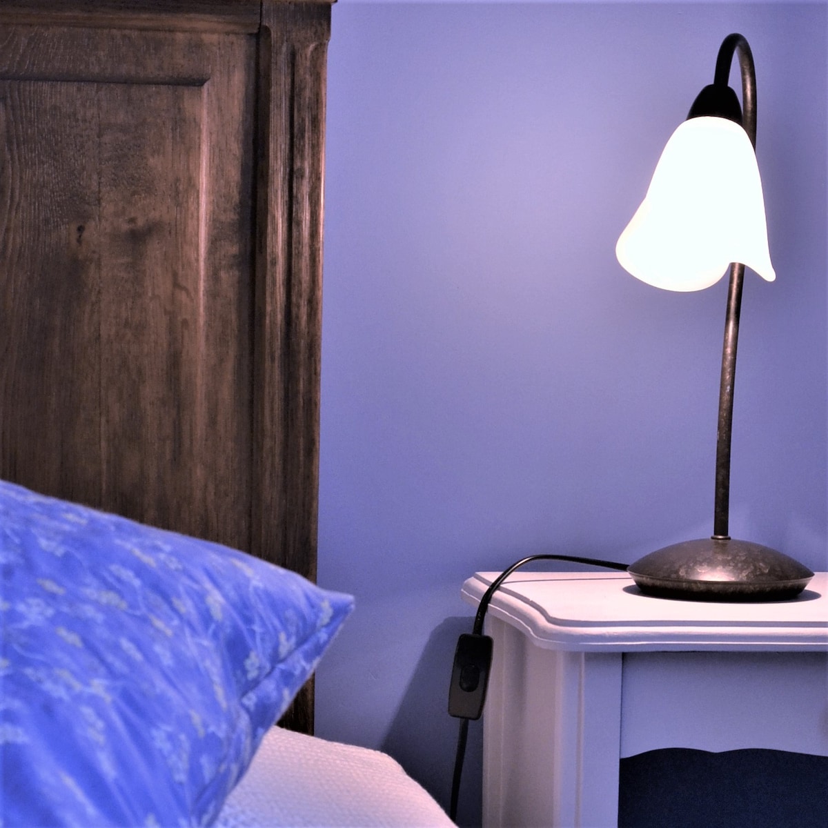 A bedside table is adorned with a softly glowing lamp, featuring a curved neck and a frosted shade. A blue pillow rests against a wooden headboard, complementing the calming blue wall behind. The cord from the lamp lies neatly along the floor.
