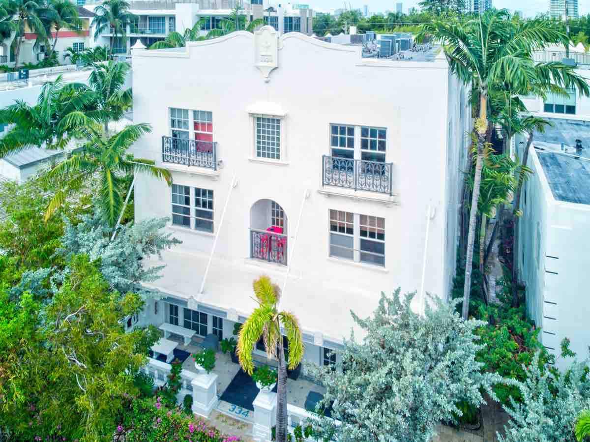 The exterior of the condominium showcases a charming white façade adorned with elegant balconies. Lush greenery frames the building, with palm trees and flowering plants enhancing the serene atmosphere of the peaceful neighborhood.