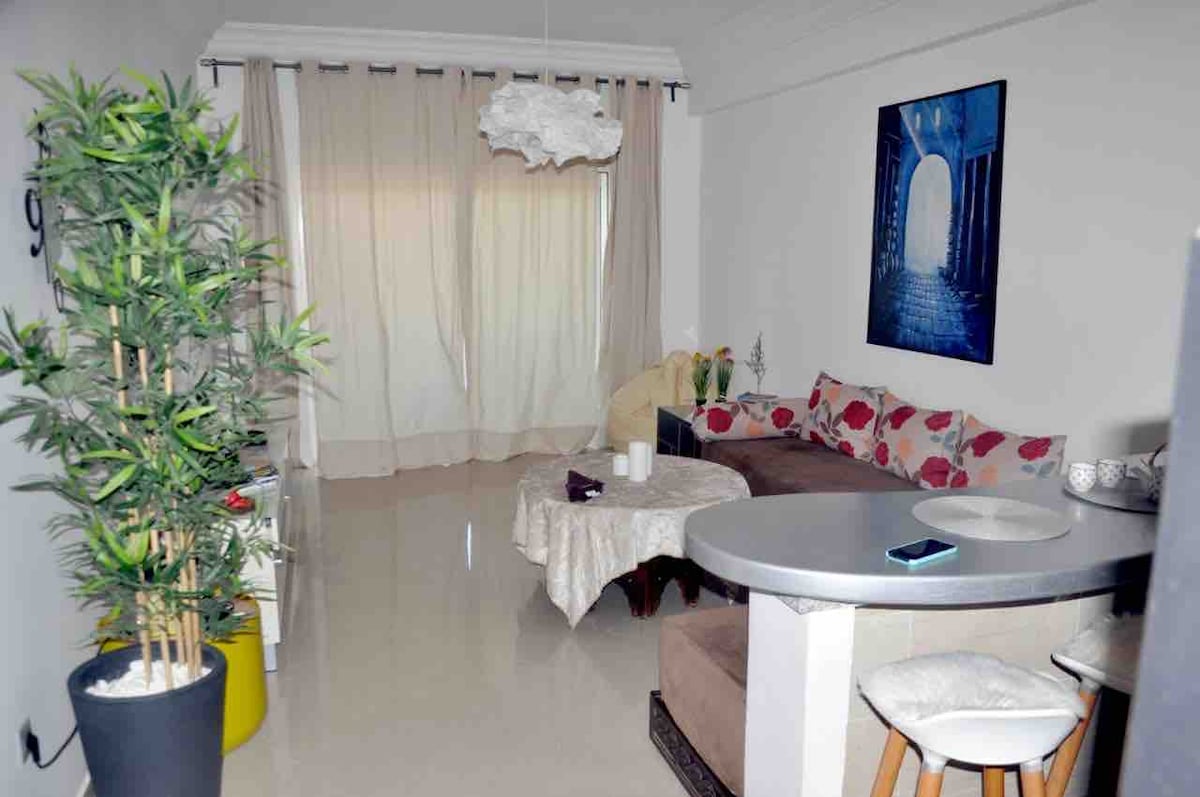 A living area features a curved sofa adorned with floral patterns, positioned near sheer drapes that allow soft light to filter in. A round table sits in the foreground with candles, while a tall plant adds a touch of greenery to the space.
