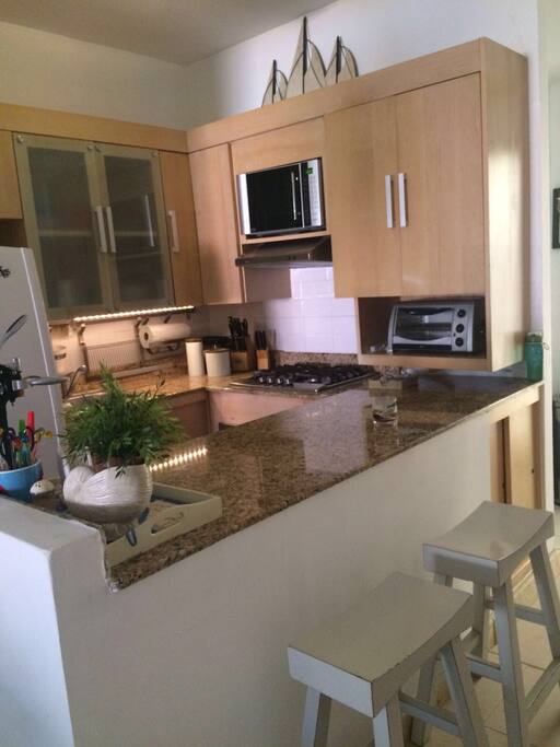 Partial panoramic view of the kitchen with stove,electric oven,sink,couple of seats next to the laundry area and entrance.