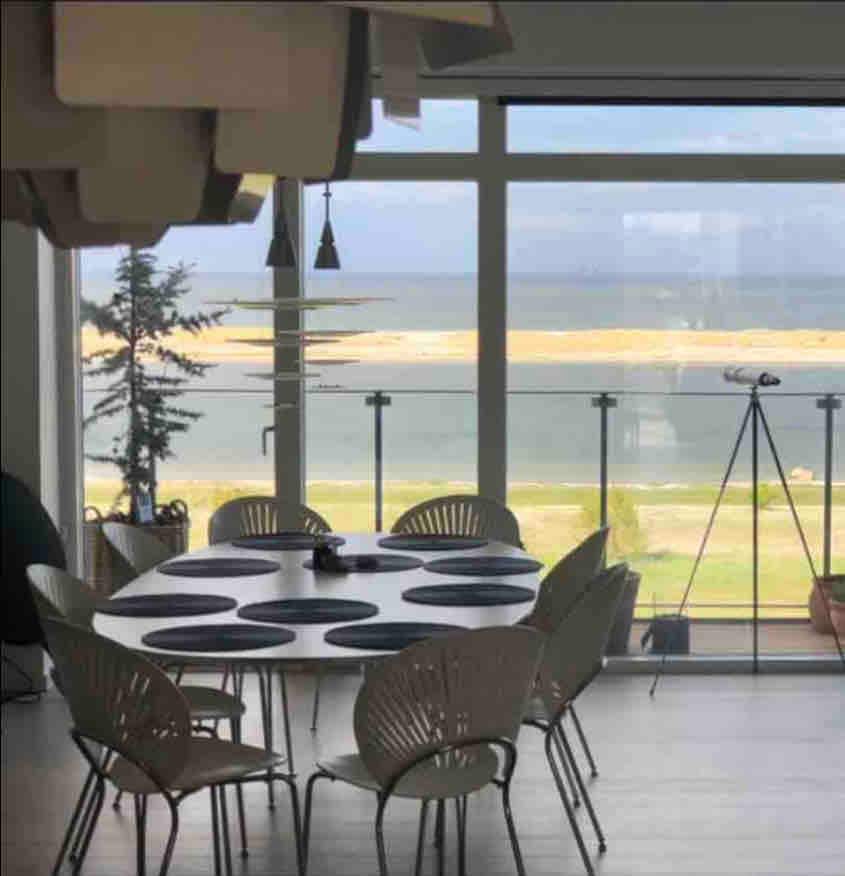 A dining area features a round table set with placemats, surrounded by several modern chairs. Large windows offer a view of the waterfront, with natural light illuminating the space. Minimalist decor enhances the open feel of the room, which reflects a calm and airy environment.