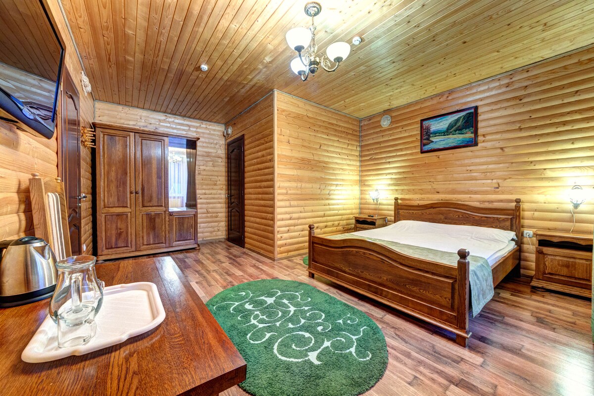 A spacious room features wooden walls and a ceiling, complemented by warm lighting. A large bed with a light green bedspread sits against the wall. A wooden wardrobe and a TV are visible, along with a cozy area rug beneath the bed.