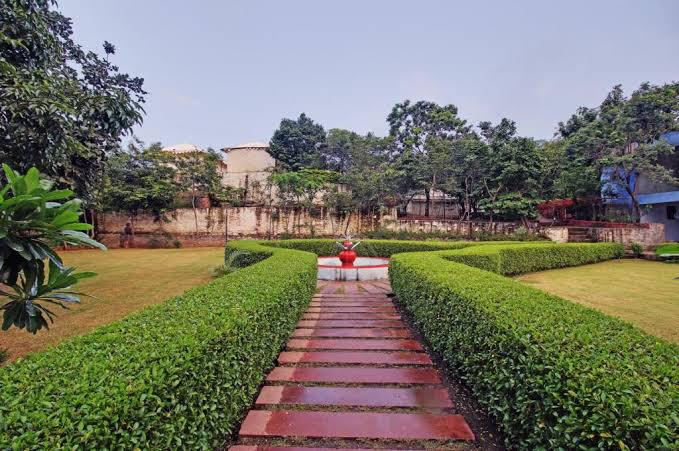 A well-maintained garden path lined with trimmed hedges leads to a central fountain. The smooth stones create a clear walkway through the lush greenery, with trees and low shrubs framing the area. The serene atmosphere is enhanced by open sky views in the background.