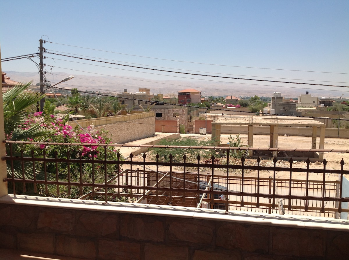 A view from the villa's balcony captures a rural landscape with scattered buildings and blooming flowers in the foreground. The backdrop features distant hills under a clear blue sky, providing a glimpse of the peaceful surroundings.
