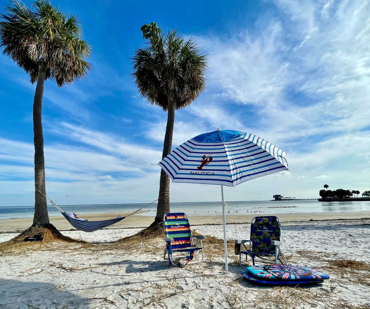 A serene beach scene is presented with two palm trees flanking a sandy area. Colorful beach chairs are positioned beside a striped umbrella, providing shade. In the background, a calm ocean and distant pier are visible under a bright blue sky with scattered clouds.