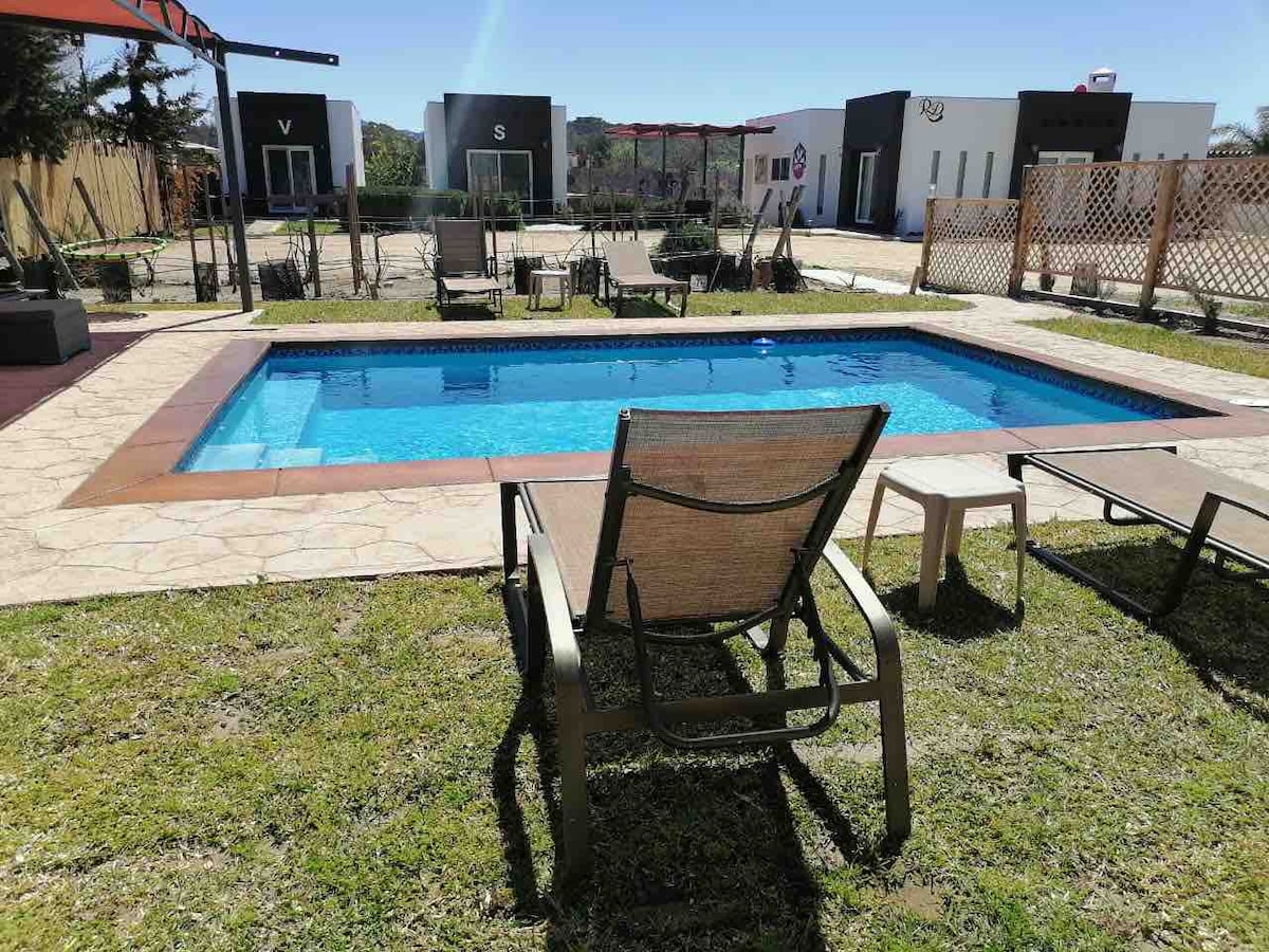 A crystal-clear swimming pool is surrounded by manicured grass and modern chaise lounges. The area is finished with stone paving and features several shaded spots, providing a relaxed setting to unwind, with minimalist structures visible in the background.