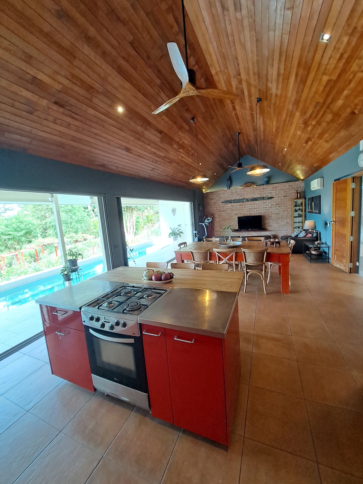 An open-plan kitchen and dining area features a modern red cabinetry and stainless steel appliances. The high wood-paneled ceiling enhances the spaciousness, while large glass doors provide views of the pool and garden, creating a bright, airy atmosphere.