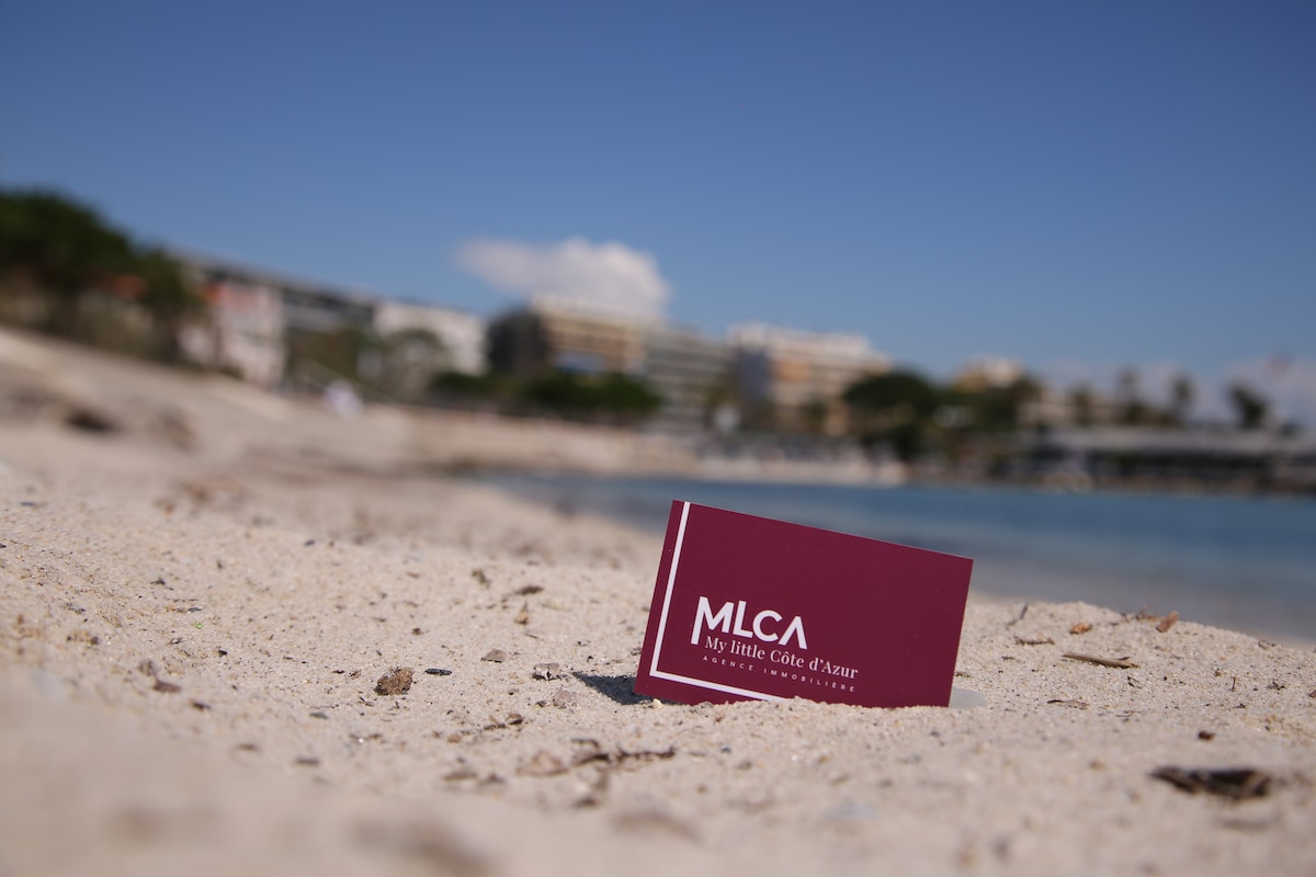 A branded card is positioned on light-colored sand at the beach. The calm water and distant shoreline are visible in the background, suggesting a coastal environment with buildings and greenery set against a clear blue sky.