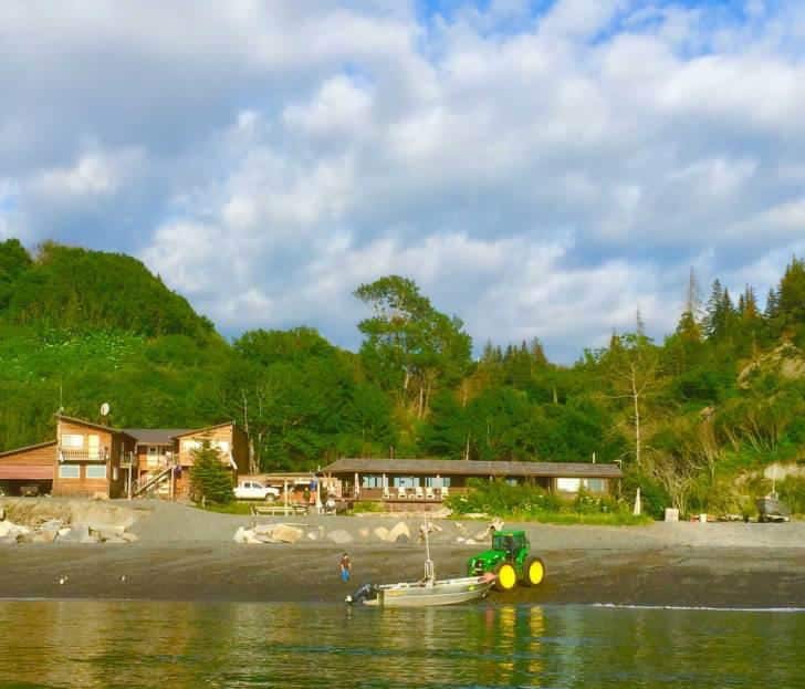 A beachfront view features a wooden lodge set against a backdrop of lush green hills. A small boat is being launched from the shore, accompanied by a tractor. The sky is partly cloudy, adding depth to the scenic coastal landscape.
