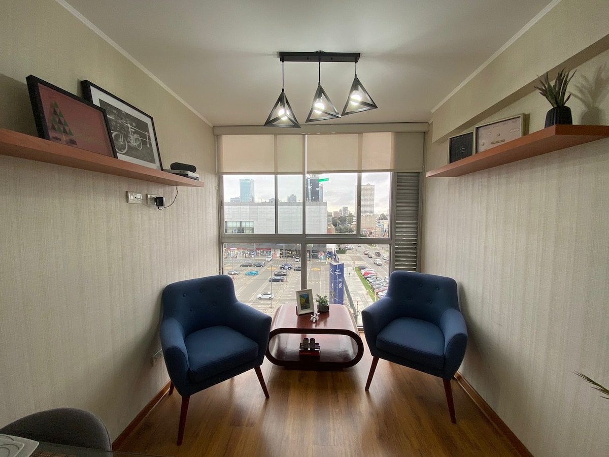 A seating area is illuminated by a modern light fixture. Two blue chairs are positioned opposite each other around a wooden coffee table. Large windows offer a view of the cityscape, allowing natural light to fill the space, enhanced by neutral-toned walls and wooden flooring.
