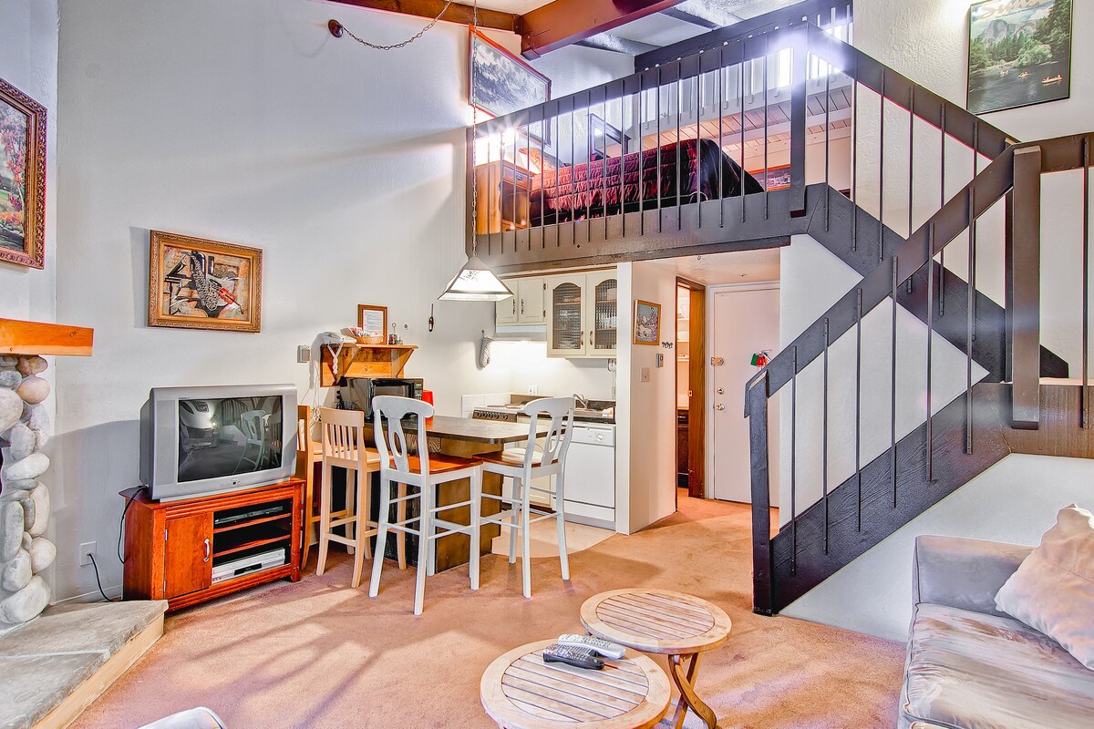 A spacious living area is visible, featuring a sofa, coffee table, and a television on a wooden stand. A dining table with chairs is adjacent to an equipped kitchenette. A staircase leads to a lofted sleeping area, with a bed partially visible above.