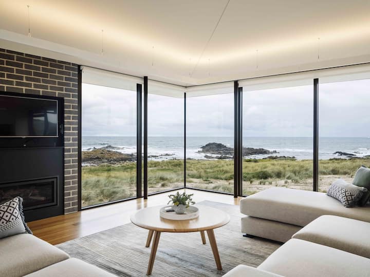  The image shows a stunning coastal living room with floor-to-ceiling windows offering panoramic views of the ocean and rugged shoreline. The space features a neutral-toned lounge area, a minimalist coffee table, and a sleek fireplace built into a brick wall. The open layout allows natural light to flood the room, enhancing the calming and serene atmosphere. The combination of modern design and the breathtaking coastal landscape creates an ideal retreat for relaxation and enjoying the beauty of nature.