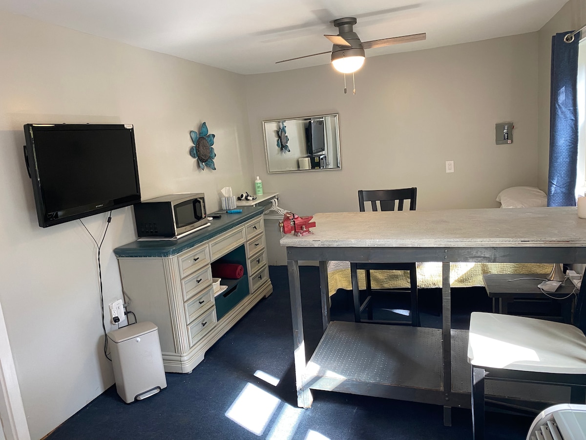 A spacious room is featured, containing a large table surrounded by chairs. A flat-screen TV and a microwave are mounted on a dresser. Natural light enters through a window, illuminating the gray walls and dark blue carpet. A ceiling fan adds a modern touch to the decor.