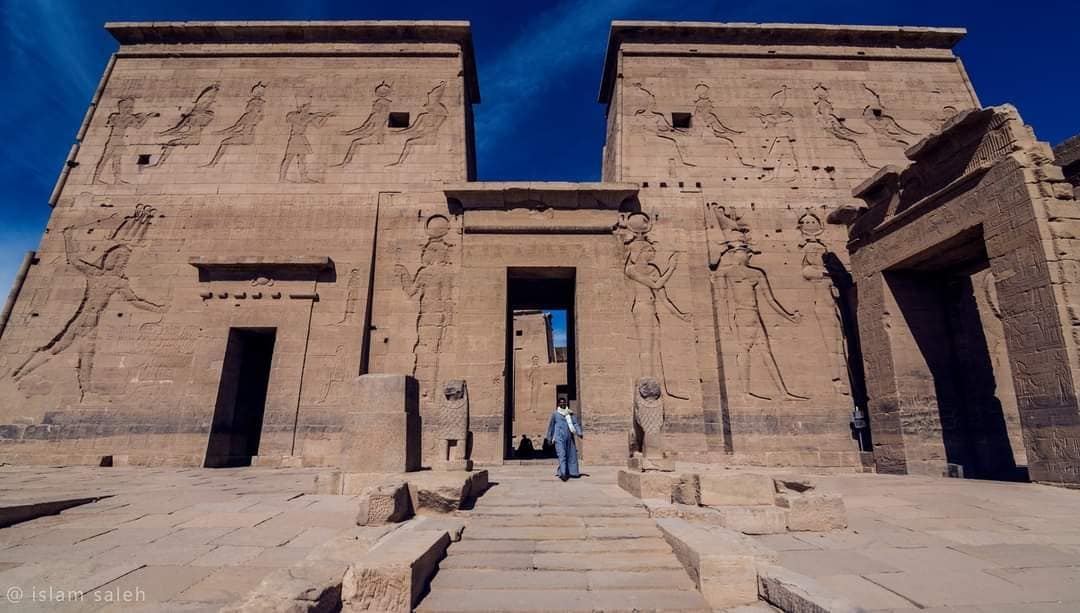 The image depicts the grand entrance of a historic temple, featuring intricately carved stone walls adorned with detailed hieroglyphics. Clear blue skies complement the monumental structure, which appears expansive, with a wide pathway leading toward the centered doorways.