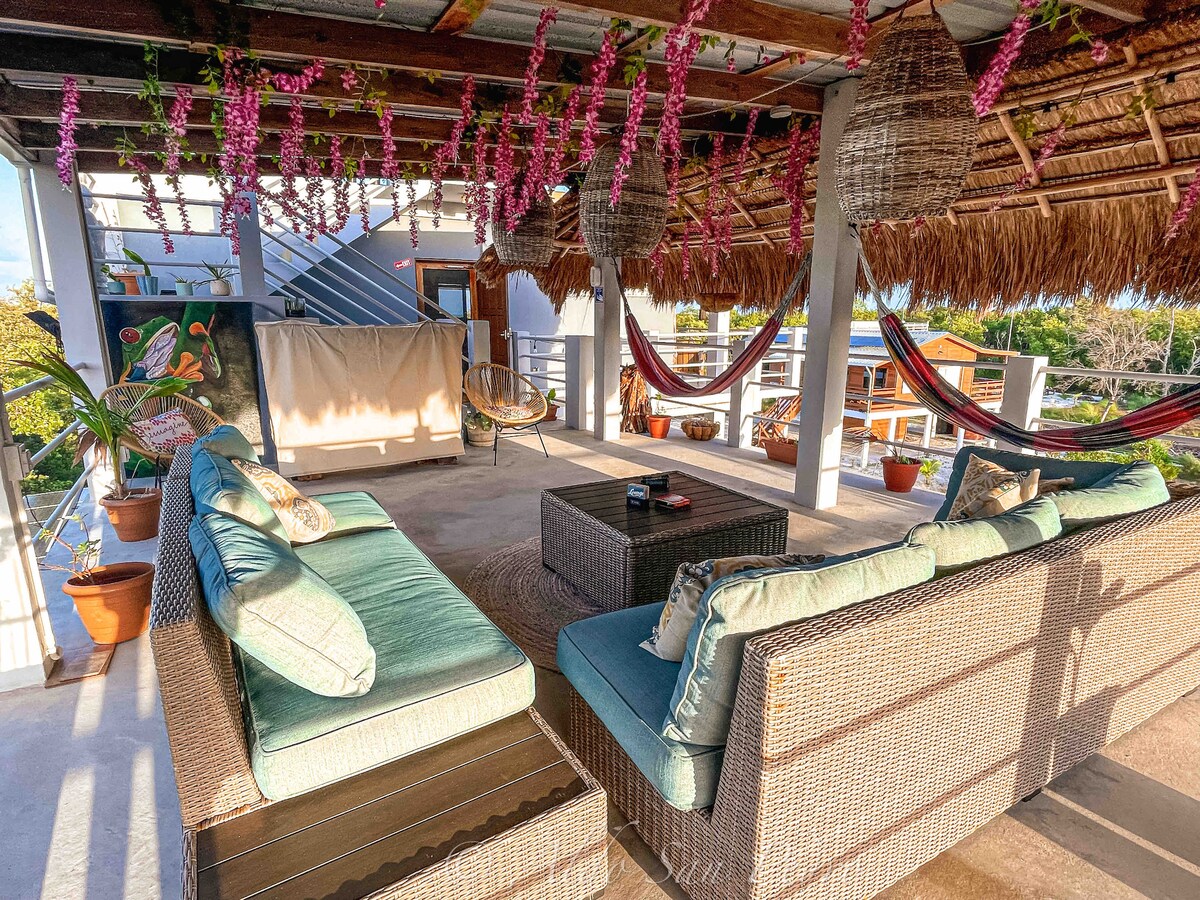 A rooftop lounge area features comfortable sectional seating adorned with soft cushions. Hammocks hang from the ceiling alongside woven light fixtures and hanging plants, creating a relaxed atmosphere. The open space is designed for gatherings while providing scenic views of the surrounding mangrove forest.