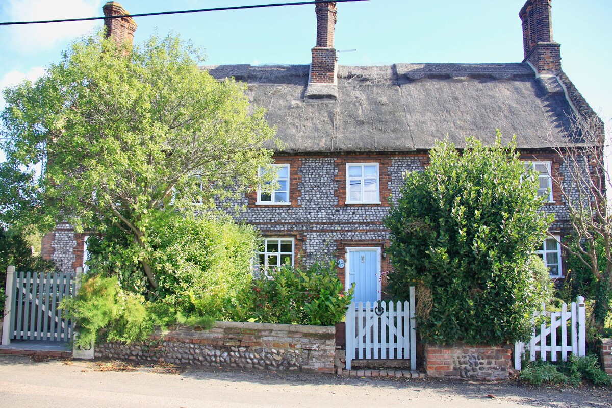 A charming Norfolk flint cottage with a thatched roof is surrounded by lush greenery. A white picket fence encloses the front garden, which features a variety of plants and small trees, creating an inviting entrance to the home.