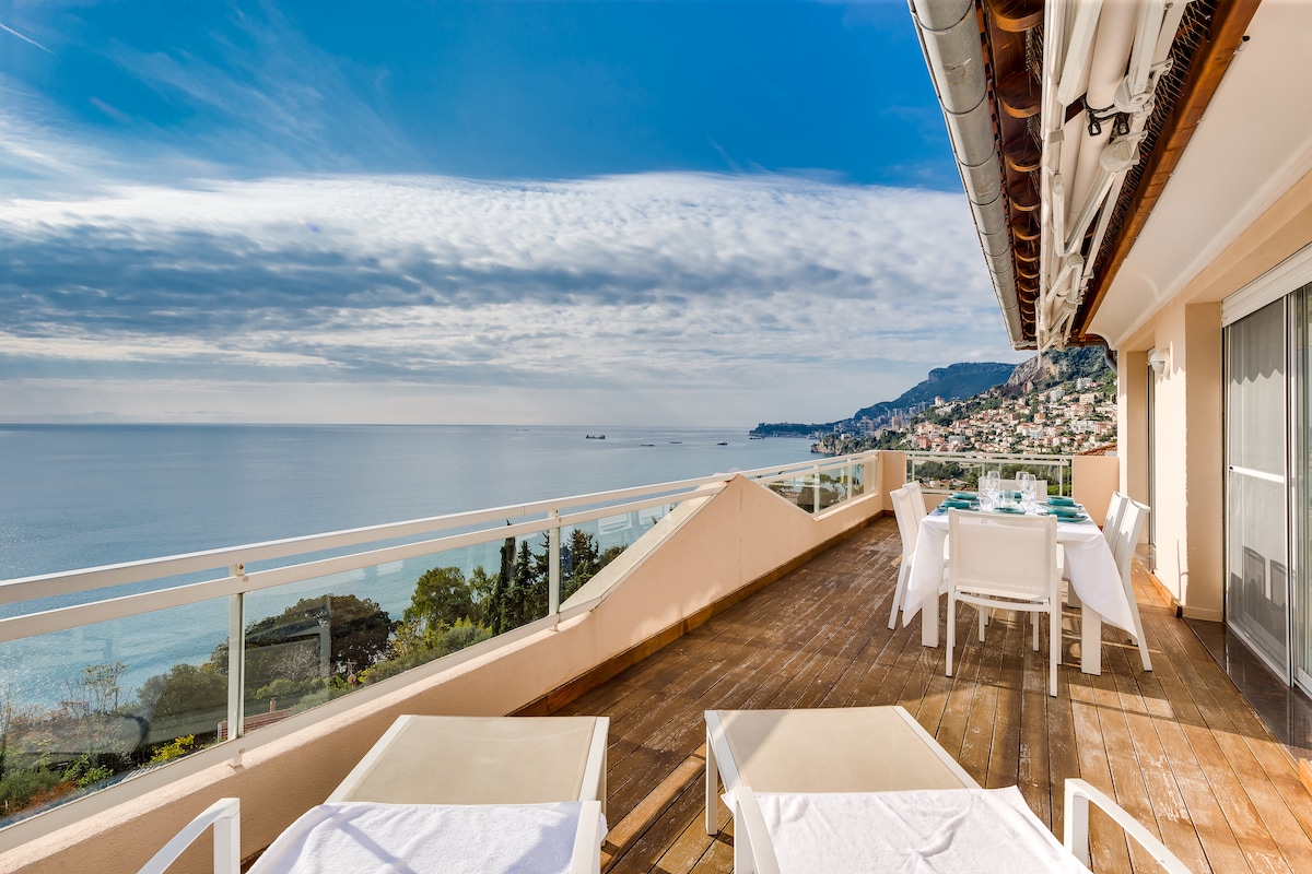 A spacious terrace overlooks the tranquil sea, showcasing a stunning panoramic view of the coastline. Several sun loungers are positioned on the wooden deck, accompanied by a dining table set for meals. The sky features soft clouds, accentuating the serene atmosphere.