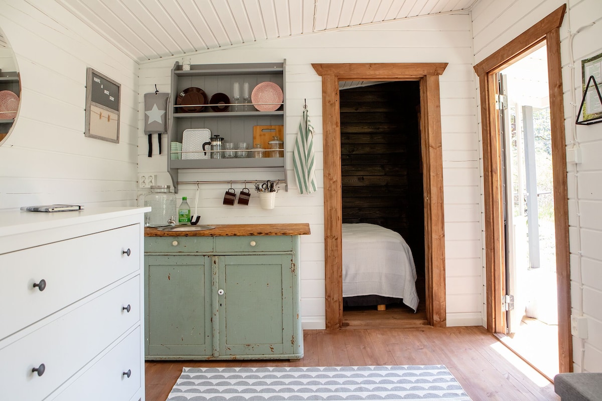 An inviting interior space is displayed, featuring a practical kitchenette with shelves containing dishes and utensils. A rustic wooden table complements a white dresser, while an open door leads to a cozy sleeping area. Natural light brightens the room through the nearby entrance.