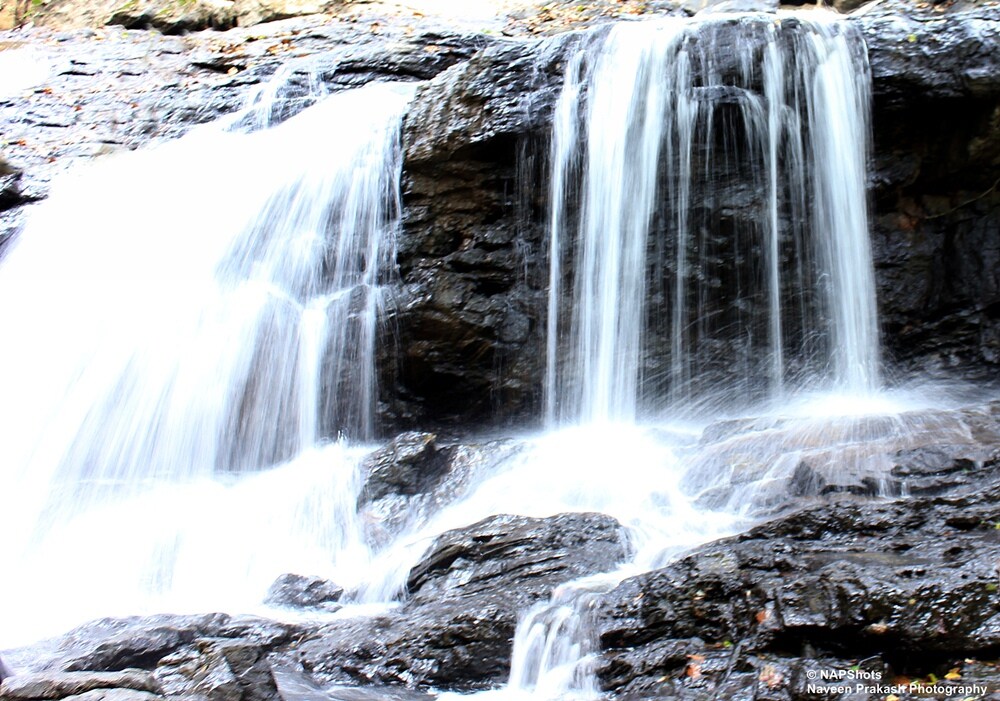 A cascading waterfall flows gracefully over dark, textured rocks. The water creates a gentle mist, adding a soft quality to the rugged landscape. Surrounding vegetation may enhance the natural setting, contributing to a serene and tranquil ambiance.