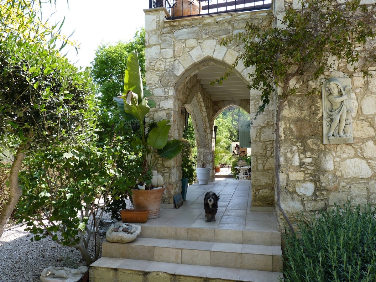 A stone pathway leads through a charming archway, bordered by lush greenery and potted plants. The natural light highlights the textured surface of the stone facade. A small dog is seen standing on the steps, adding a warm presence to the outdoor space.