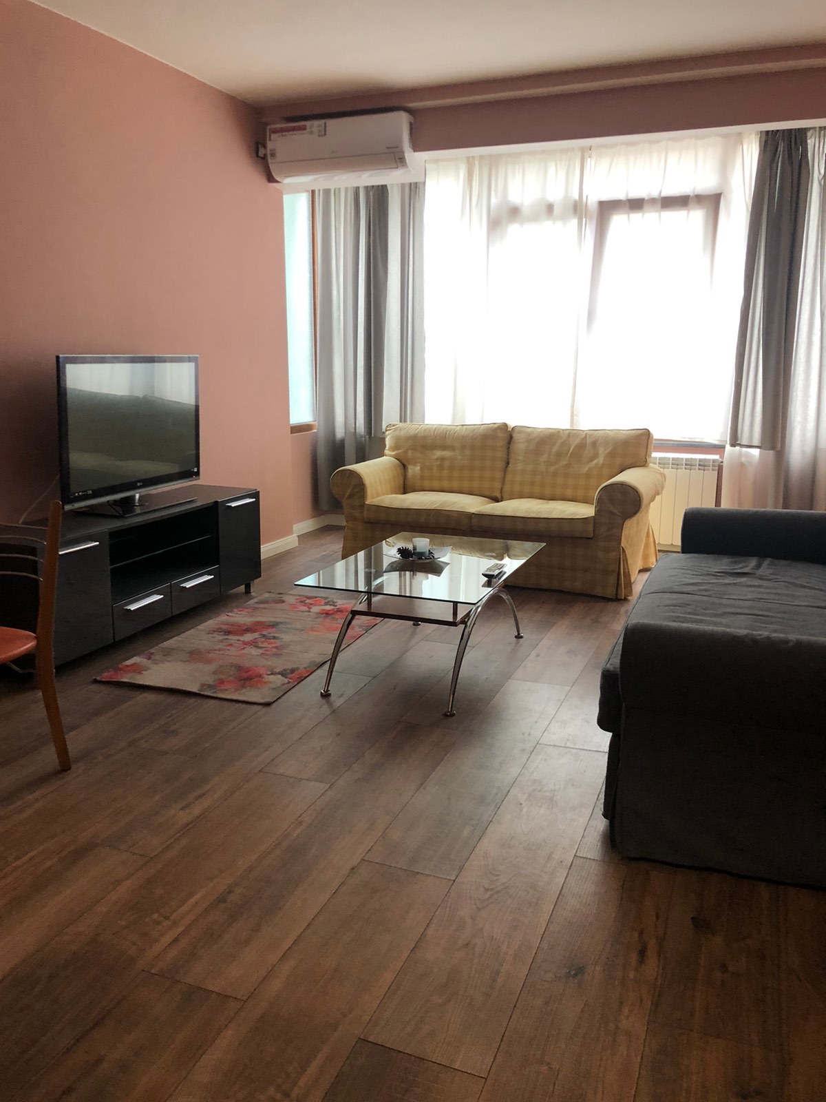 A well-lit living area features a comfortable yellow sofa and a dark-colored couch, complemented by a glass coffee table in the center. A television is positioned on a black media unit. Light streams in through large windows, framed by soft curtains.