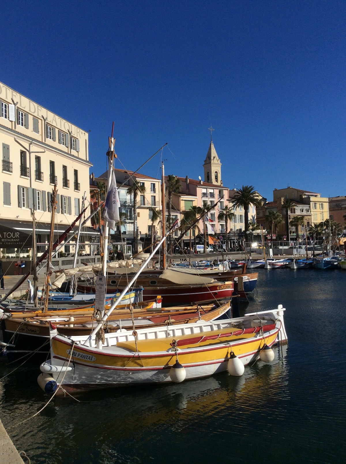Fishing boats with colorful hulls are moored in a calm harbor, surrounded by buildings featuring pastel facades and palm trees. A tall clock tower rises in the background, while clear blue skies illuminate the scene, creating a serene waterfront atmosphere.