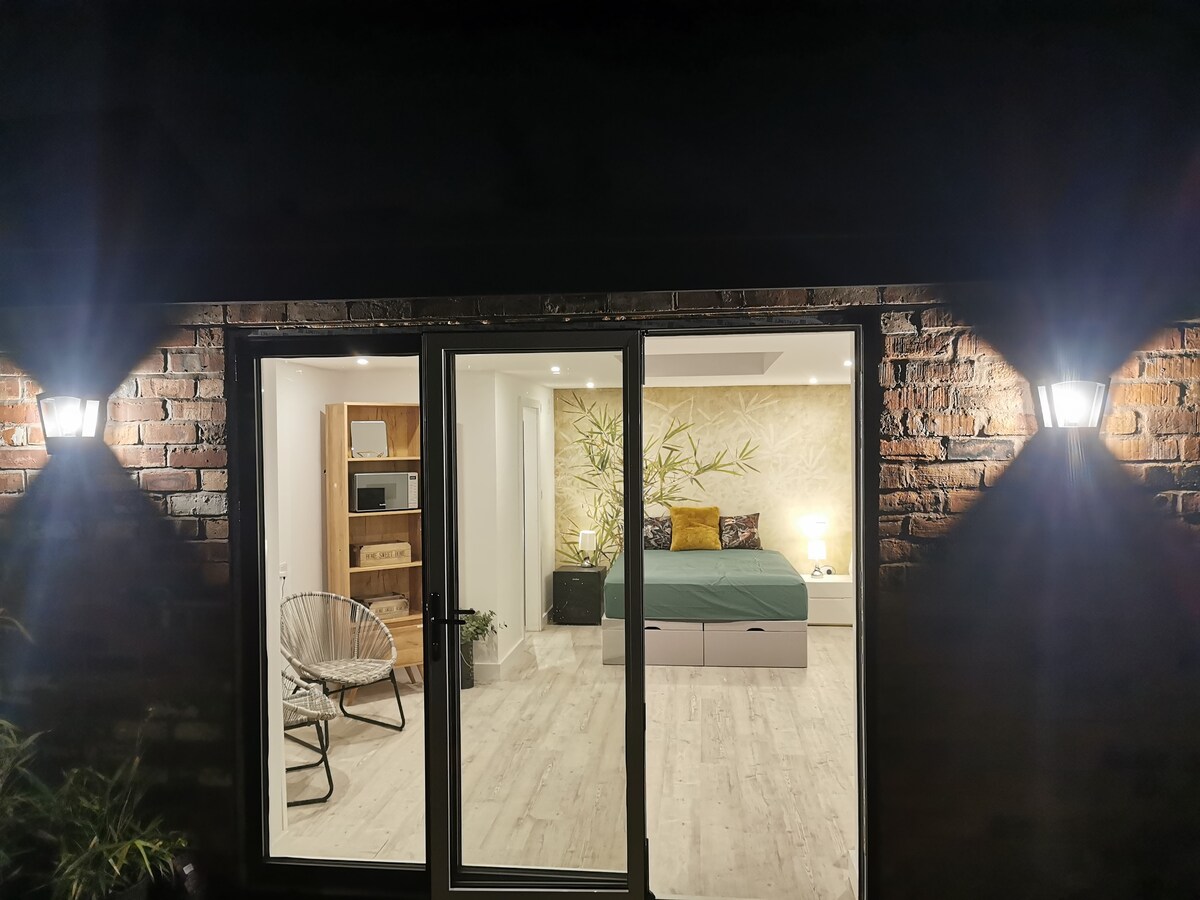 The exterior view shows a cozy private annex illuminated by exterior lights. Large glass doors provide a glimpse of the interior, which features a spacious bed against a decorative wall. Nearby, a comfortable chair and functional kitchenette are visible, enhancing the welcoming space.